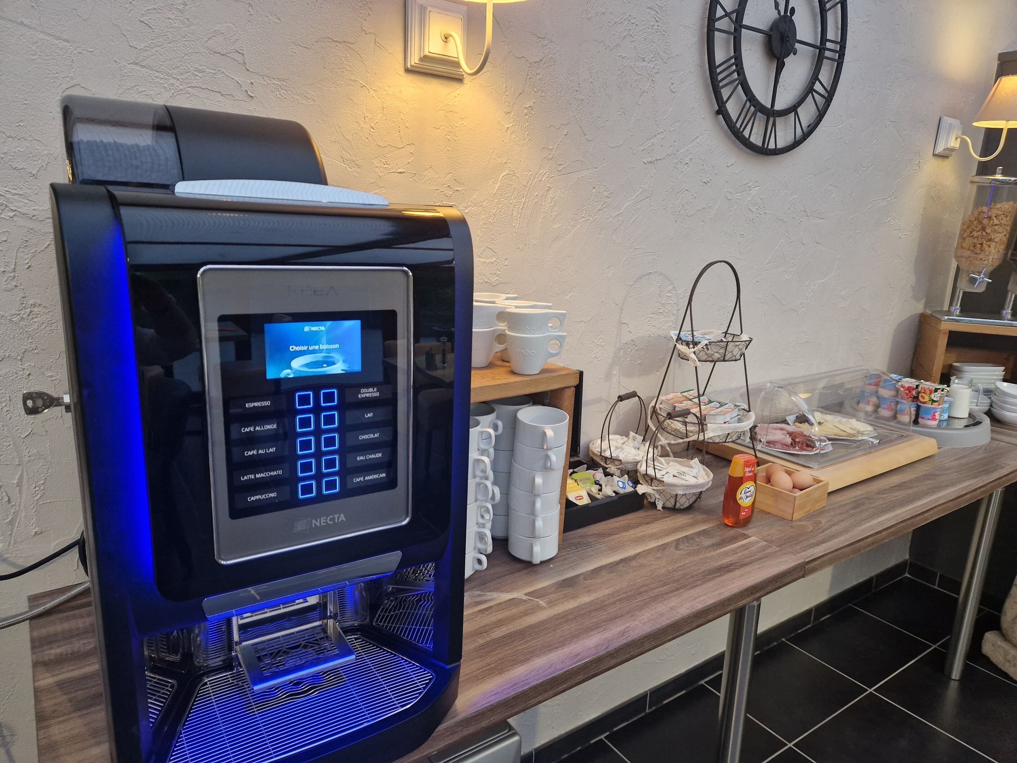 Una cafetera con pantalla digital, un reloj en la pared y una barra de desayuno con diversos productos.