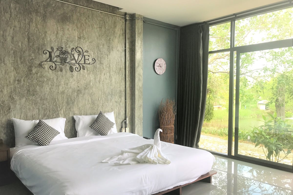 A bedroom with a large bed, a decorative wall piece, a clock, and a view of greenery outside.