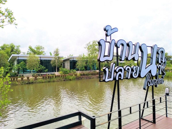 A sign with Thai script, a modern building with glass windows, and a water body with a wooden dock. - Baannok Plaina Homestay (Klaeng)