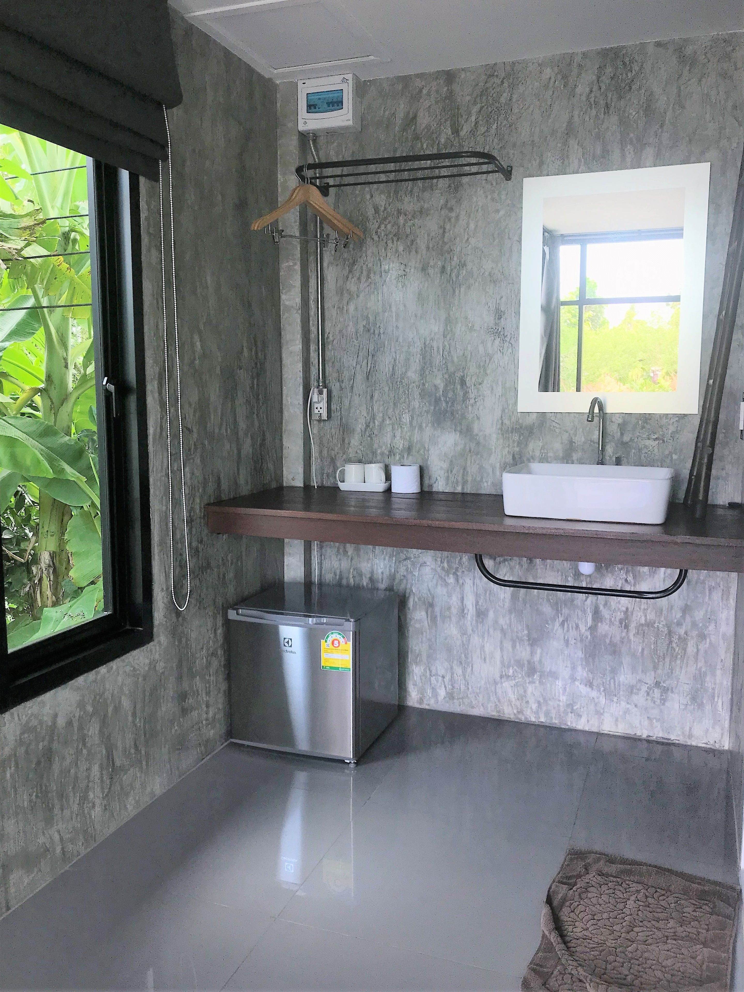 Une salle de bain moderne avec un mur en béton, un réfrigérateur en acier inoxydable, un lavabo avec un miroir et une fenêtre donnant sur un espace verdoyant.