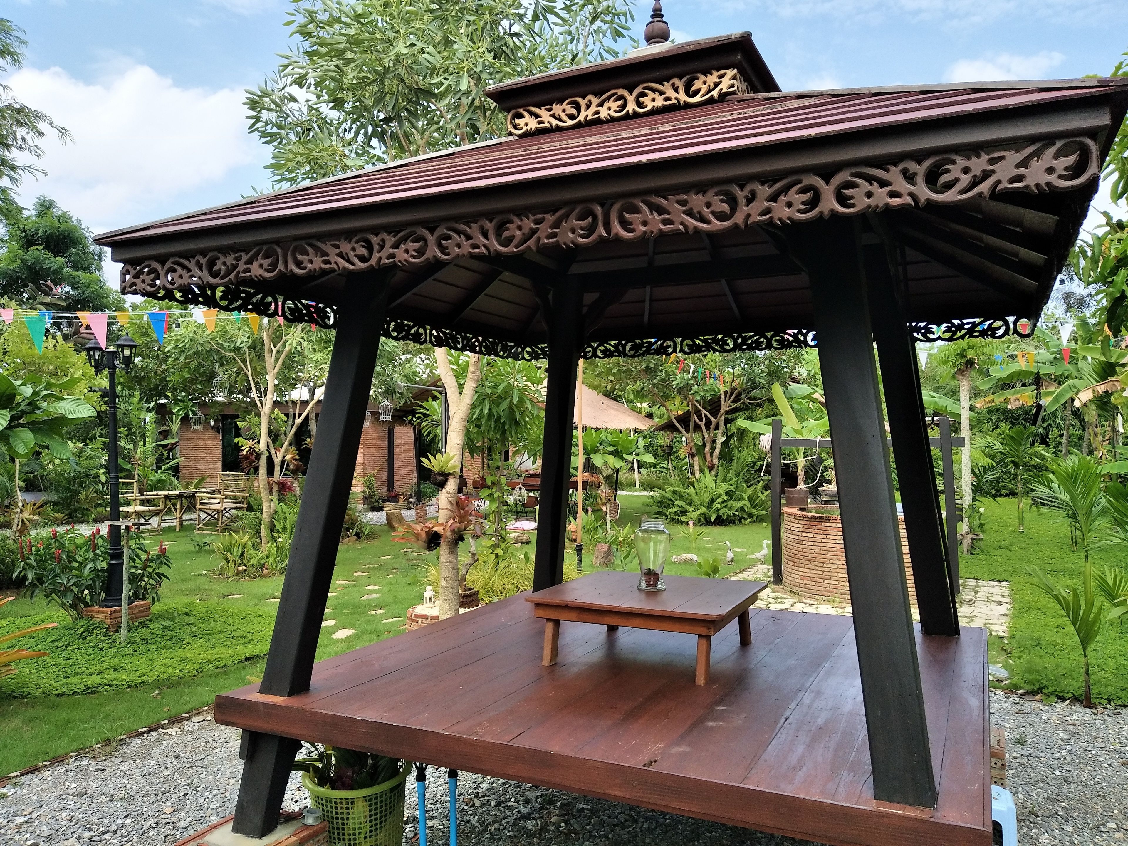 Un kiosque avec un banc en bois, entouré d’un jardin où poussent différentes plantes, et un mur de briques en arrière-plan.