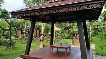 Un kiosque avec un banc en bois, entouré d’un jardin où poussent différentes plantes, et un mur de briques en arrière-plan.