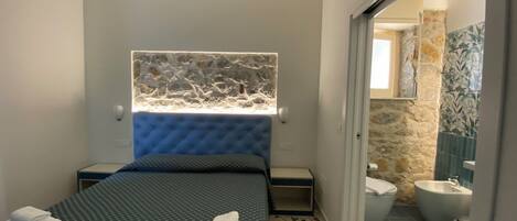 A hotel room with a bed, a bathroom visible through an open door, and a stone wall with a window above the bed.