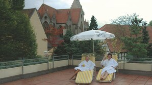 Twee mensen ontspannen op ligbedden onder een grote parasol, met een historisch gebouw op de achtergrond.