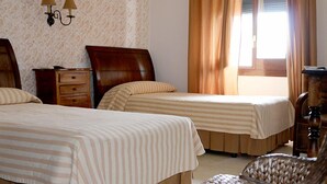 A hotel room with two beds, a wooden nightstand, a lamp, and a window with curtains. - Hostal - Restaurante Emilia (Trujillo)