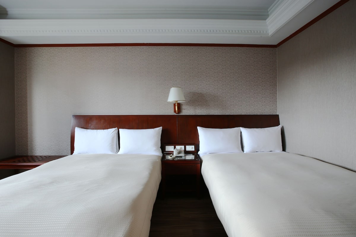 A hotel room with two single beds, a wooden headboard, a nightstand with a lamp, and a small table.