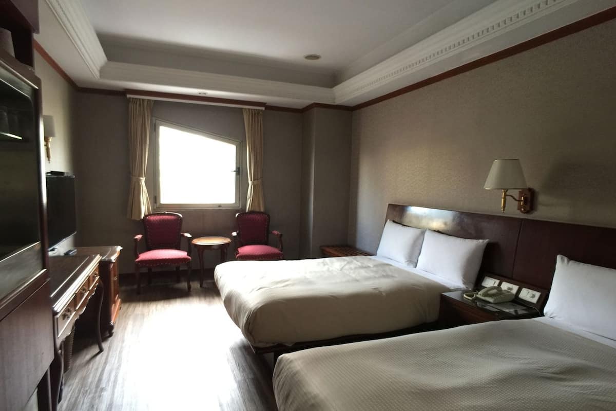 A hotel room with two beds, a desk, a chair, a TV, and a window with curtains.