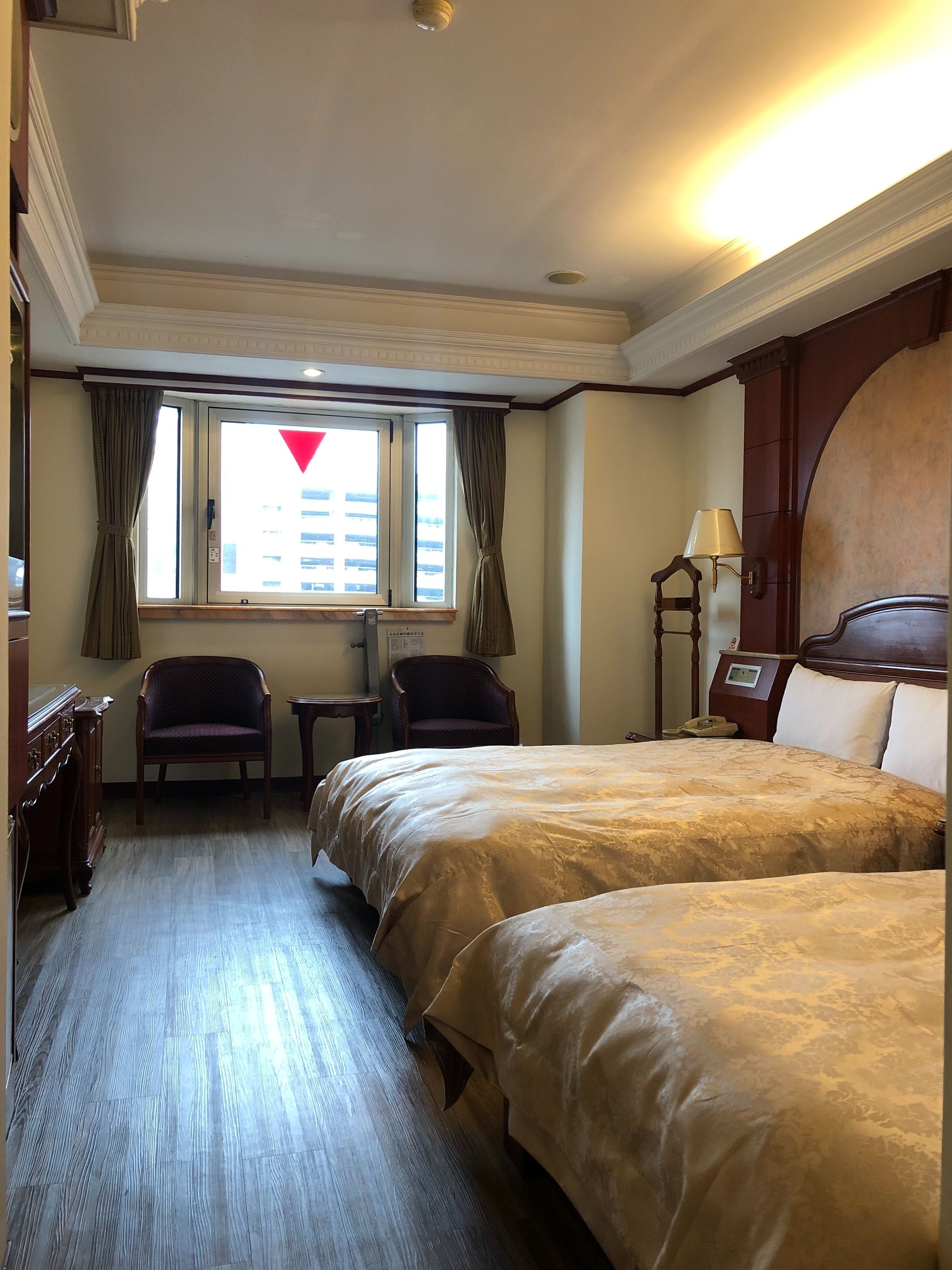 Camera d'albergo con due letti, una scrivania, una sedia, una finestra con tende e una lampada.