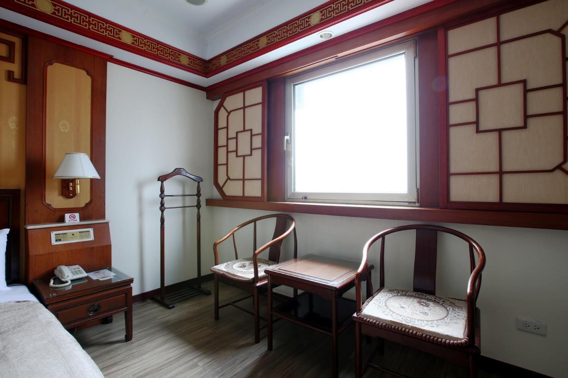 A hotel room with a traditional design, featuring a wooden desk, two chairs, a bedside table with a lamp, and a large window with a view.