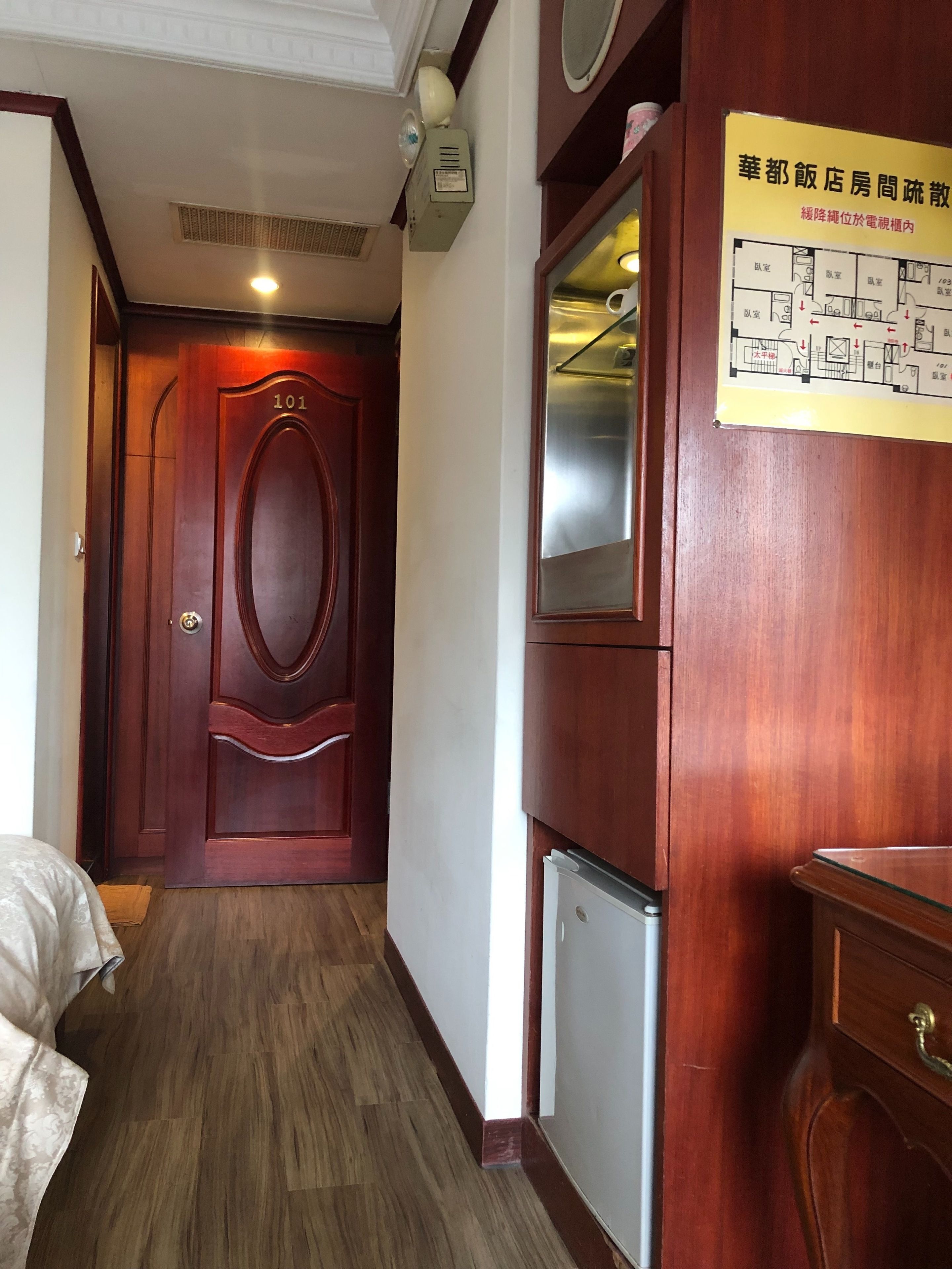 A hotel corridor with a wooden door labeled '101', a small refrigerator, and a wall-mounted mirror.