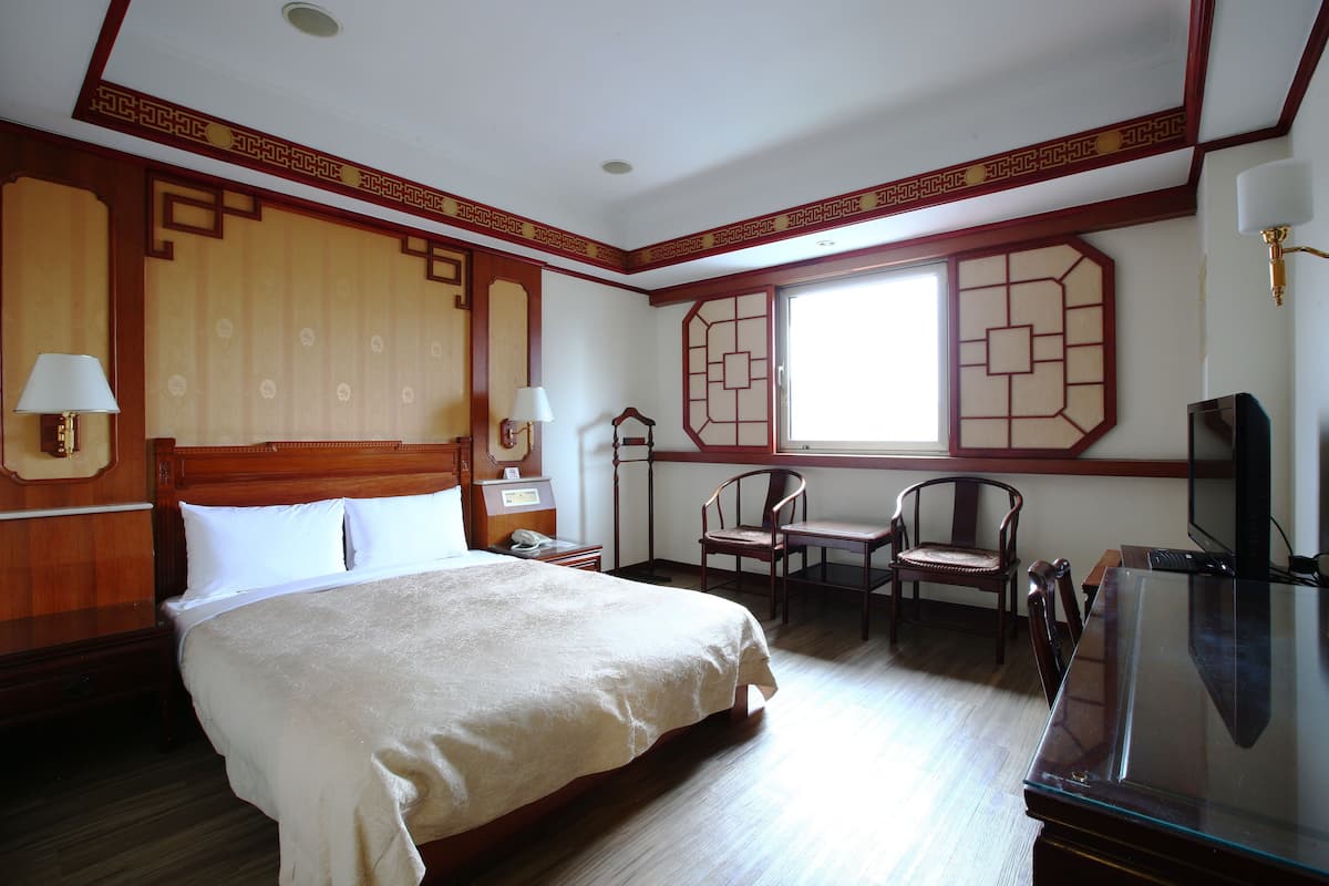 A hotel room with a large bed, wooden headboard, two chairs, a desk, and a television.