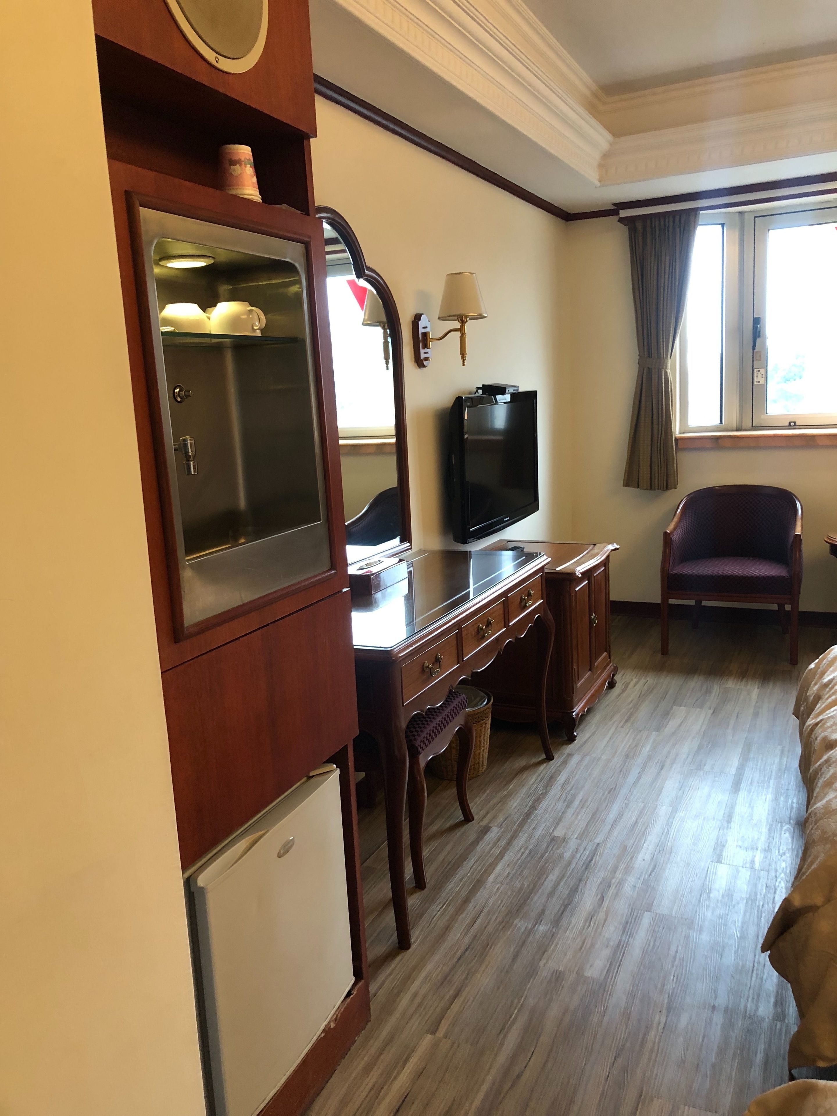 A hotel room with a wooden desk, a television, a small refrigerator, a chair, and a window with curtains.