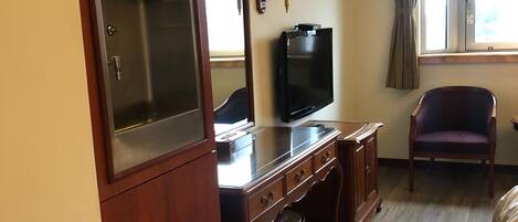 A hotel room with a wooden desk, a television, a small refrigerator, a chair, and a window with curtains.