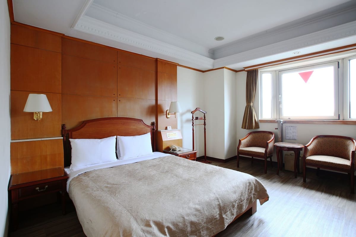 A hotel room with a large bed, a desk, two chairs, a lamp, and a window with curtains.