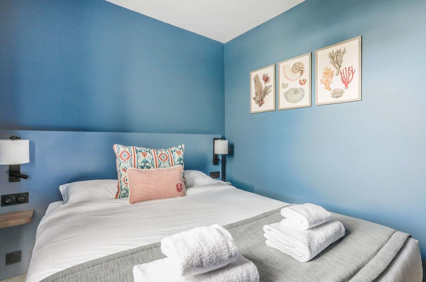 A hotel room with a blue wall, a bed with white bedding, two pillows, a bedside table with a lamp, and three framed pictures on the wall.