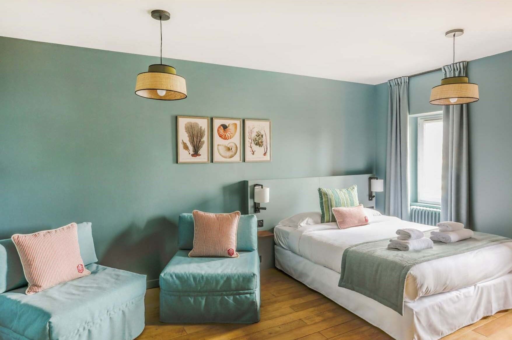 Habitación de hotel con dos camas, un sofá y una silla. Cuenta con lámparas de pared y una ventana con cortinas.