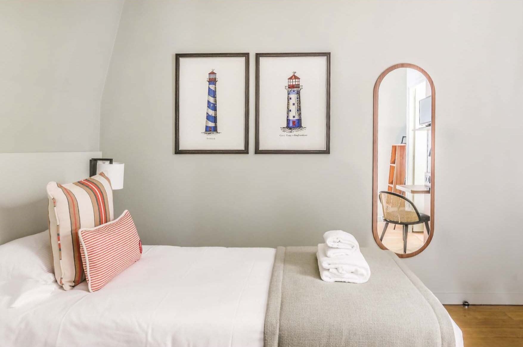 Une chambre à coucher bien rangée, avec un lit, des oreillers, un tableau encadré représentant un phare, un miroir et une chaise.