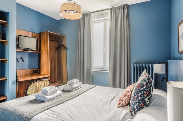 A hotel room with a bed, a TV, a wooden wardrobe, a window with curtains, and a radiator.