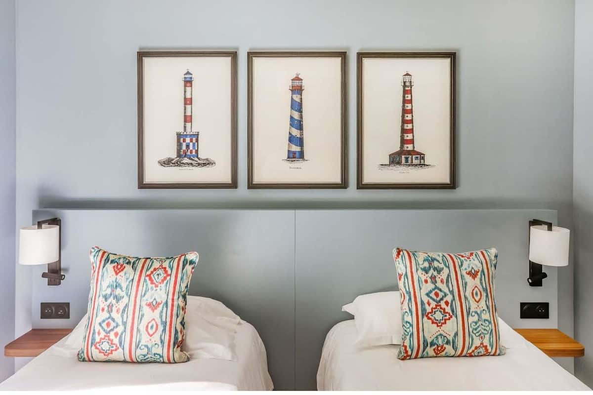 Een hotelkamer met twee bedden, elk voorzien van gekleurde kussens, een lichtblauwe muur en drie ingekaderde afbeeldingen van vuurtorens boven de bedden.