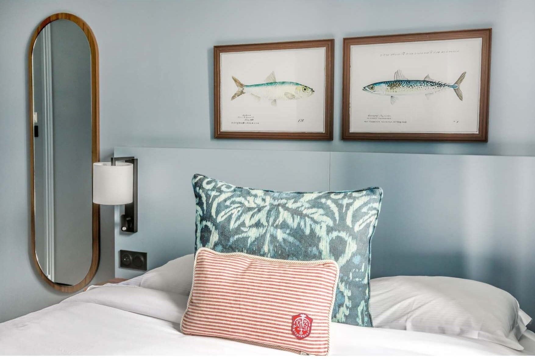 A hotel room with a bed, a large mirror, two framed fish illustrations on the wall, and a patterned throw pillow.