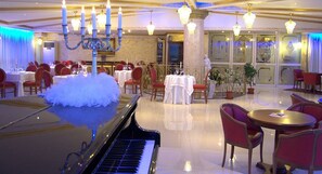 Een statige eetzaal met een piano, kroonluchters en rode stoelen.