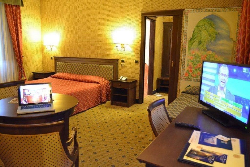 A hotel room with a bed, a desk with a laptop, a television, and a painting on the wall.