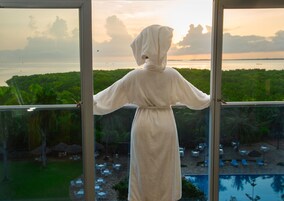 A person in a robe standing by a window with a view of a pool and outdoor seating area.