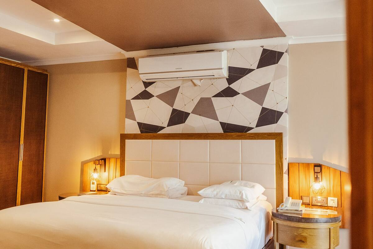 Una habitación de hotel moderna con una cama grande, suelo de madera, pared con diseño geométrico y una mesita de noche pequeña con lámpara.