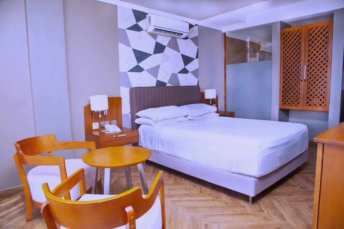 Habitación de hotel con cama, dos sillas de madera, una mesita redonda, un mesillón con lámpara y un armario con puerta persiana.