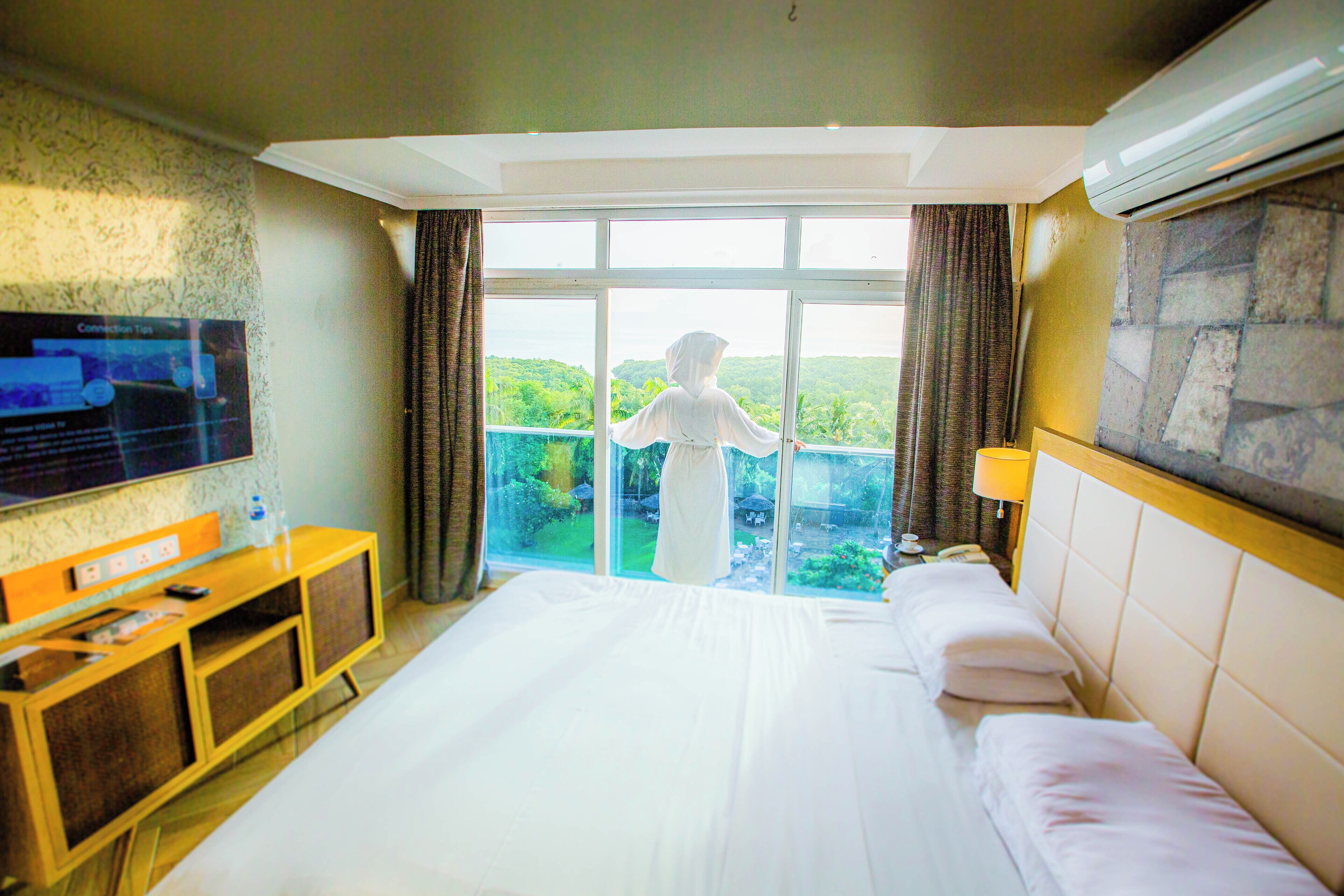 A hotel room with a large bed, a television, a wooden side table, and a view of greenery through the window.