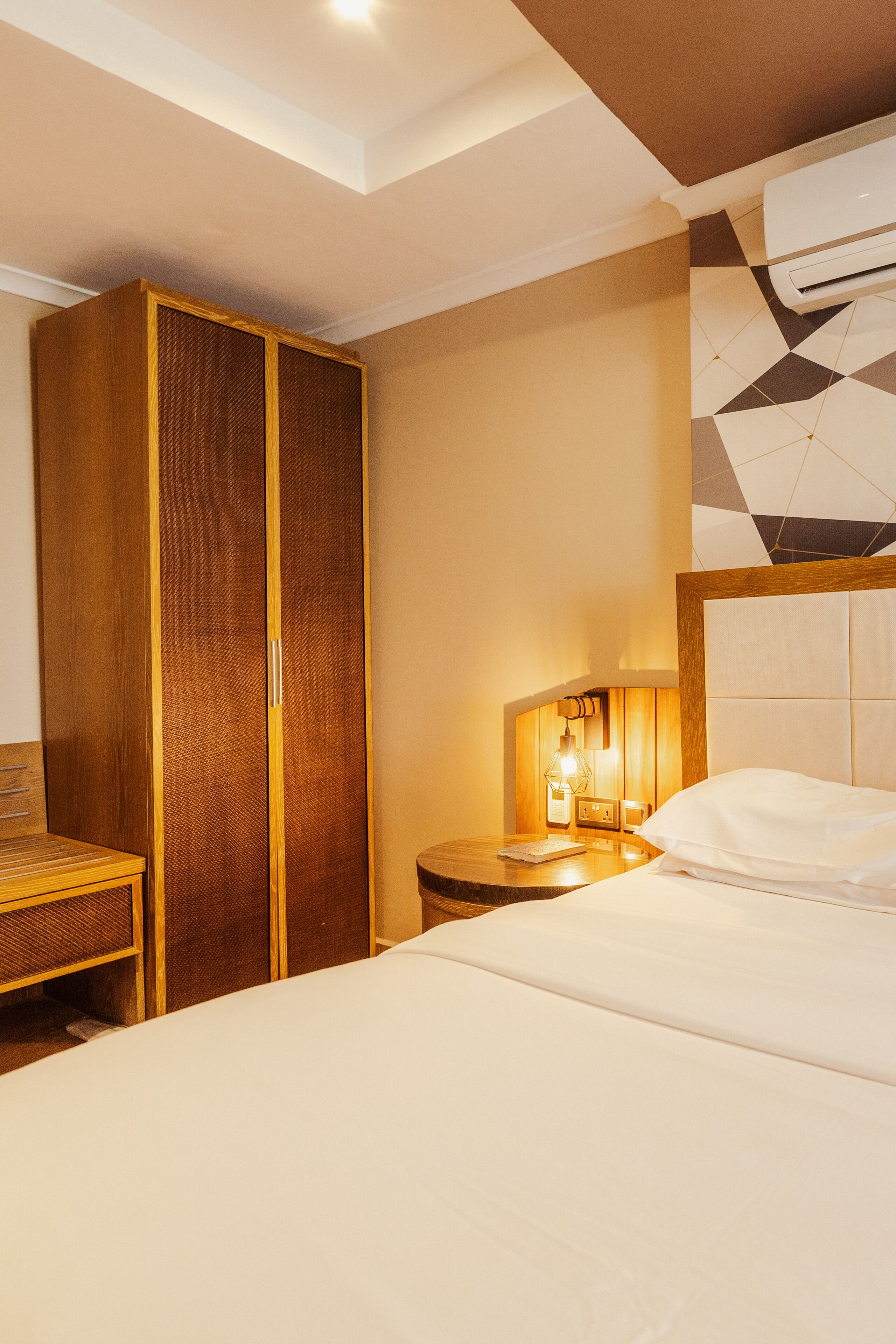 Habitación de hotel con cama, armario de madera, mesita de noche con lámpara y pared con estampado geométrico.