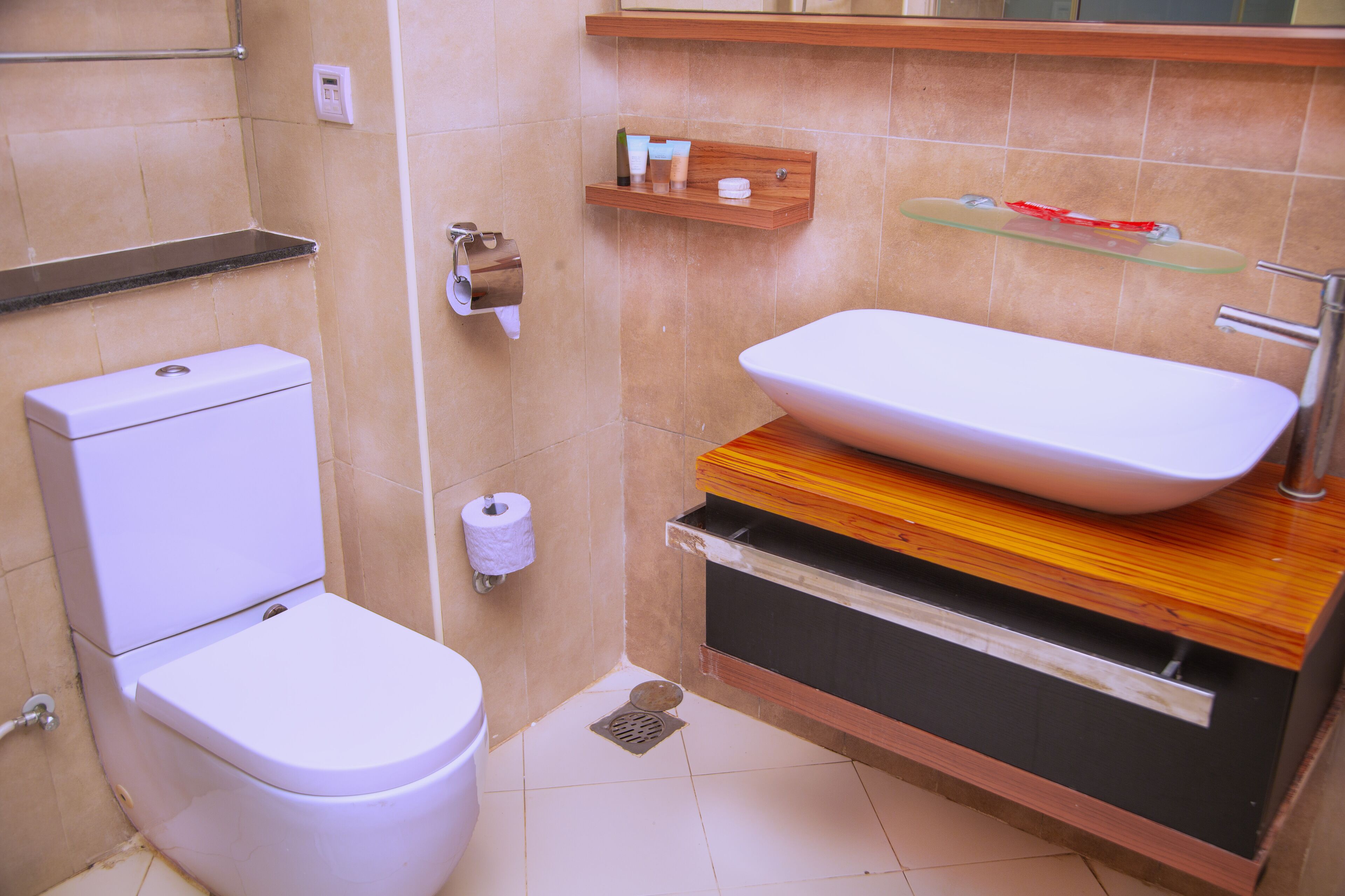 A bathroom with a toilet, a sink with a vessel basin, a wooden vanity, and a shelf with toiletries.