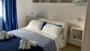 A hotel room with a bed, a bedside table, a shelf with decorative items, and a hanging rack with towels.
