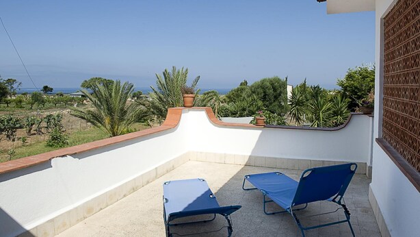 A balcony with two blue lounge chairs, a white wall, and a view of the sea and greenery.