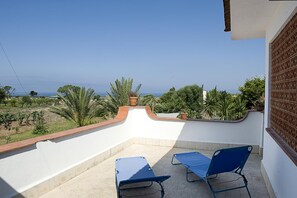 Un balcón con dos sillones azules, una pared blanca y vistas al mar y la vegetación.