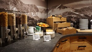 A breakfast buffet with labeled containers of yogurt and muesli, a bowl of fruit, and a wooden box of bread. - Hotel Garni Zugspitz (Farchant)