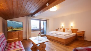 A hotel room with a wooden ceiling, a bed with white linens, a wooden nightstand, a wooden table with a vase, a checkered sofa, and a television on a wooden cabinet. - Hotel Garni Zugspitz (Farchant)