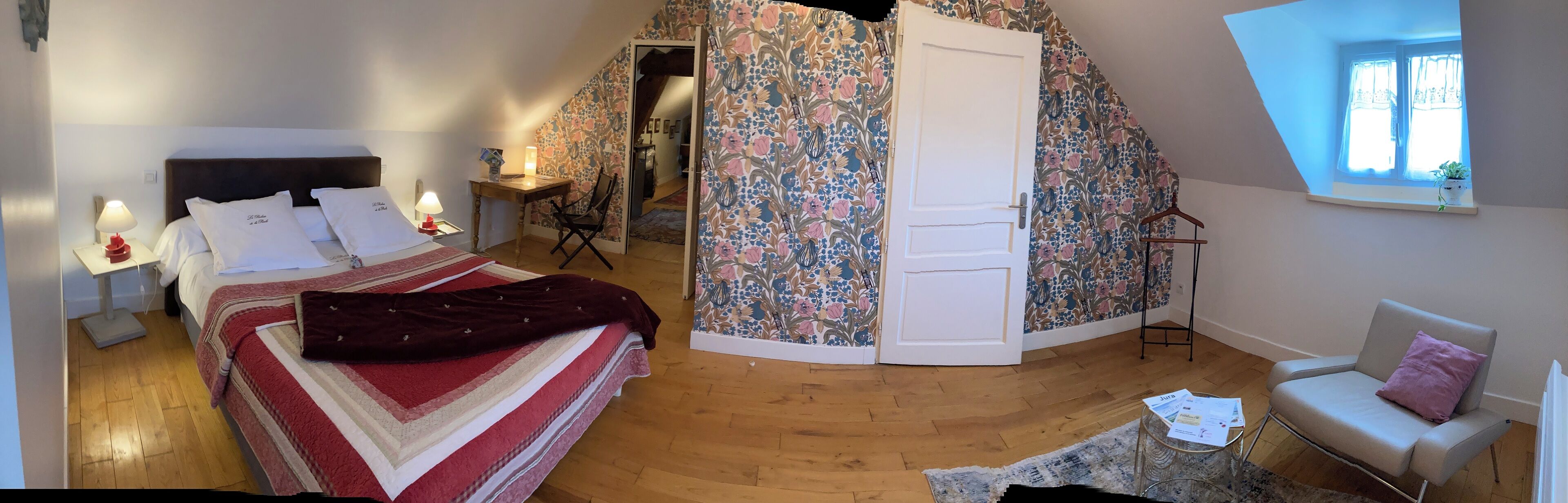 A bedroom with a bed, bedside tables, a chair, a desk, and a door leading to another room.