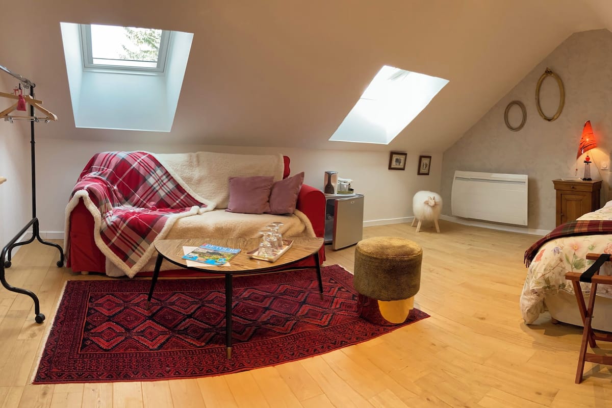 Un dormitorio acogedor en el ático con un sofá rojo y blanco, una mesita redonda pequeña, un banco de madera y una cama con ropa de cama floral. Hay dos tragaluces y un radiador en la pared.