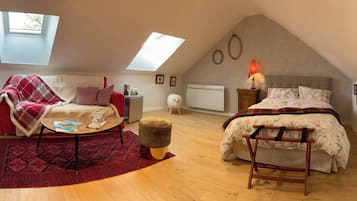 A cozy attic bedroom with a red and white sofa, a small round table, a wooden bench, and a bed with floral bedding. There are two skylights and a radiator on the wall.