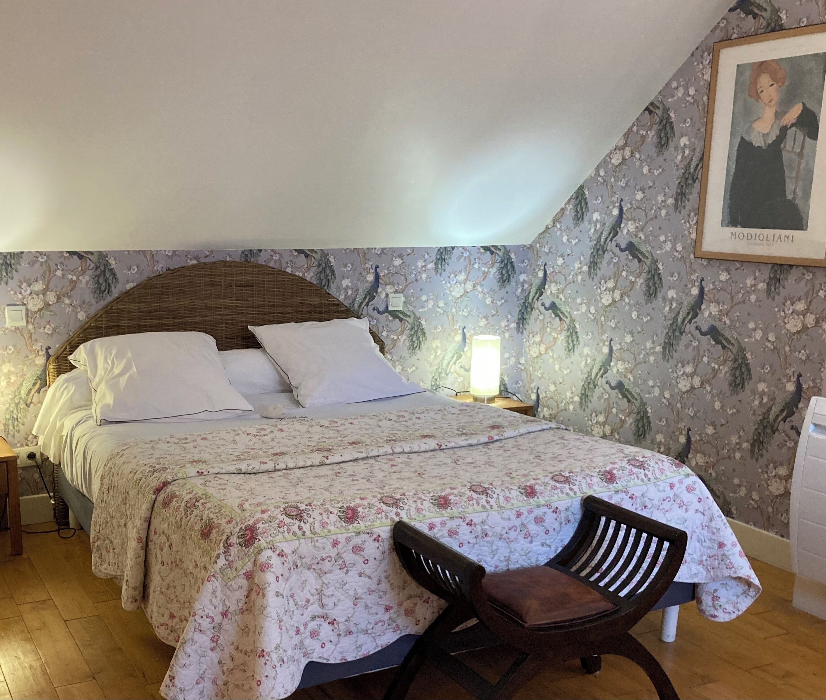 Une chambre à coucher comprenant un lit, une chaise, une table de chevet et un mur recouvert d’un papier peint à motifs floraux.