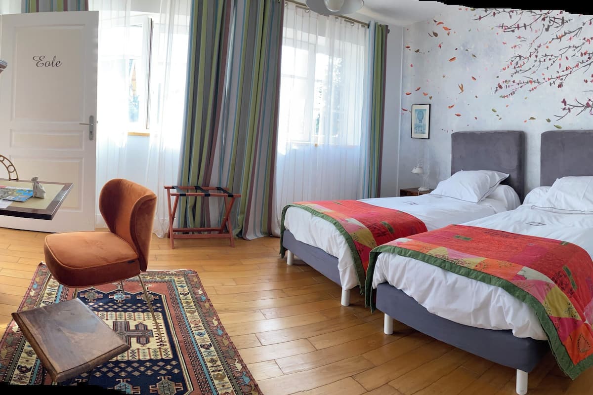 Un dormitorio con dos camas, un aparador de madera, una silla de mimbre, una mesita pequeña y una alfombra colorida.