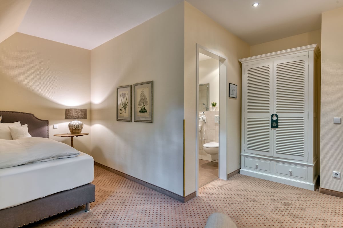 Une chambre à coucher avec un lit, une table de chevet, une lampe et des œuvres d’art accrochées au mur. On peut apercevoir une salle de bain à travers une porte ouverte.