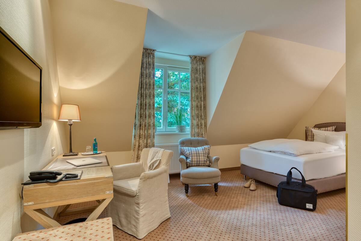 Une chambre d’hôtel avec un lit, un bureau avec une lampe, une chaise, une petite table et une fenêtre avec des rideaux.