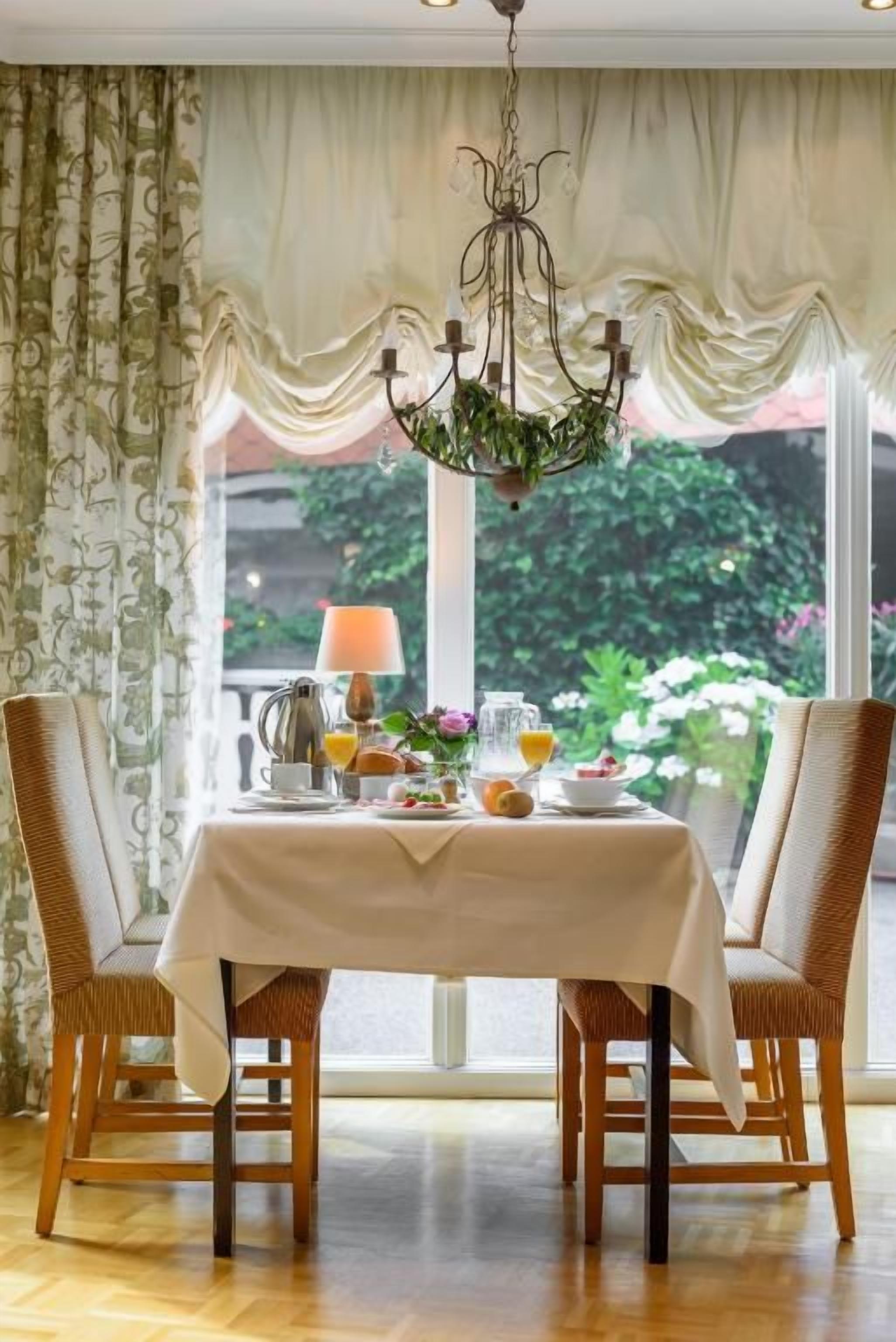 A dining area with a table set for breakfast, two wicker chairs, a chandelier, and a view of greenery outside.