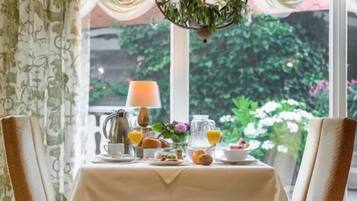 A dining area with a table set for breakfast, two wicker chairs, a chandelier, and a view of greenery outside.