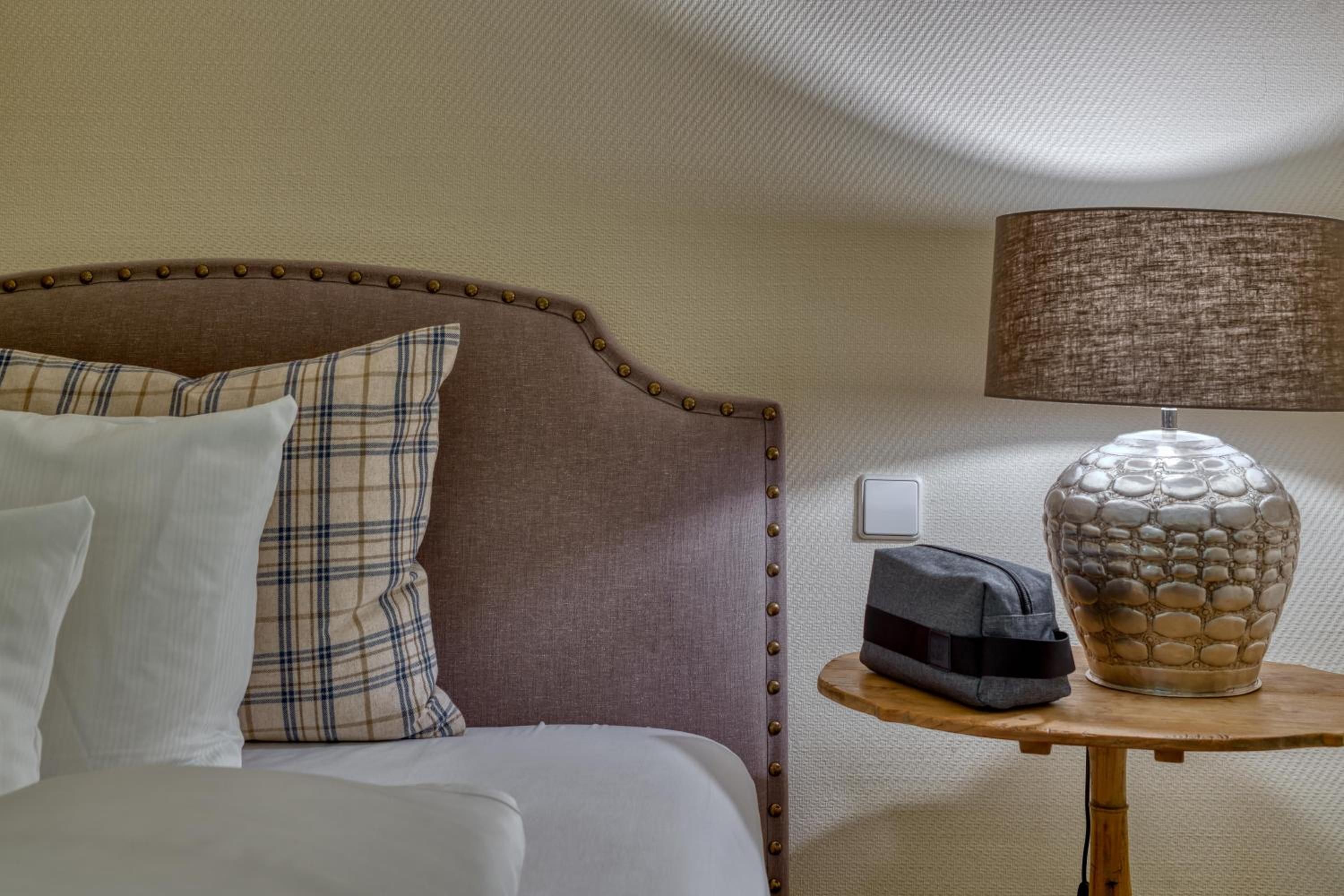 Una cama bien hecha con cabecera, una lámpara de noche y un jarrón decorativo sobre una mesita.