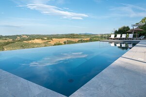 Una piscina infinita rectangular con vistas despejadas del paisaje, un área de descanso techada con sillones reclinables y una sombrilla junto a la piscina.