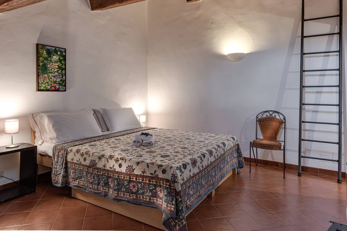 A bedroom with a bed, a nightstand, a chair, a ladder, and a wall-mounted light fixture.