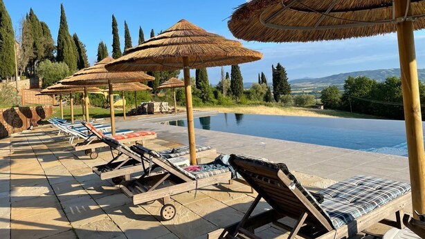 Zona piscina con lettini e ombrelloni di paglia, circondata da alberi e vista sul paesaggio.